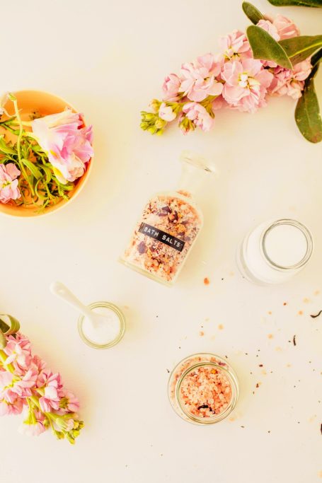 A bottle of bath salts surrounded by flowers and a small dish of salts on a light surface.